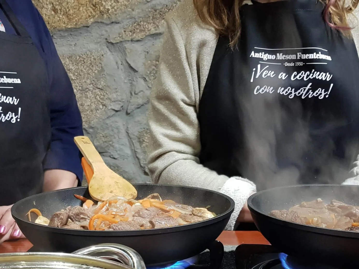 Dos mujeres con delantales negros cocinando comida en una clase de cocina.